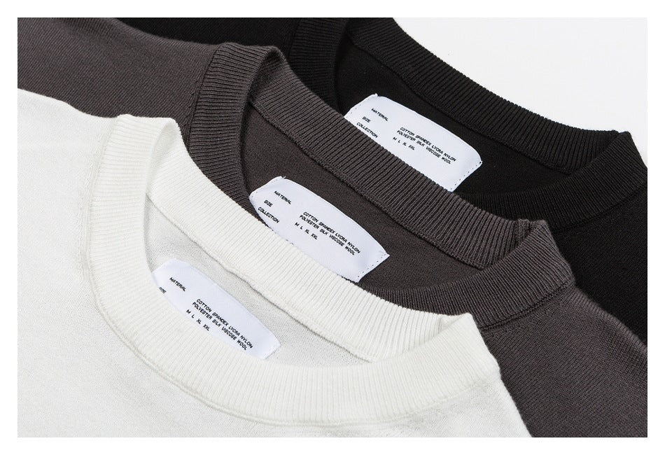 Close-up of three crew-neck knit sweaters in white, charcoal gray, and black, showing ribbed collars and inner labels.
