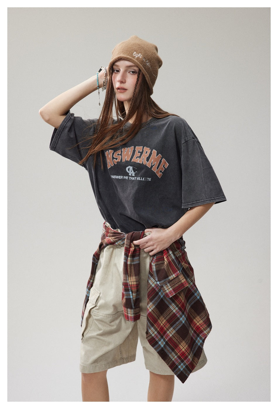 Model wearing an oversized charcoal graphic T-shirt with beige cargo shorts and a red plaid shirt tied at the waist.