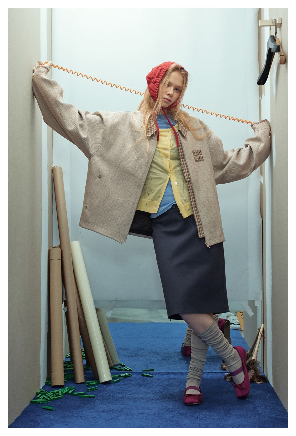 Model posing in an open taupe checked-trim jacket, layered tops and dark skirt, wearing a red bonnet.