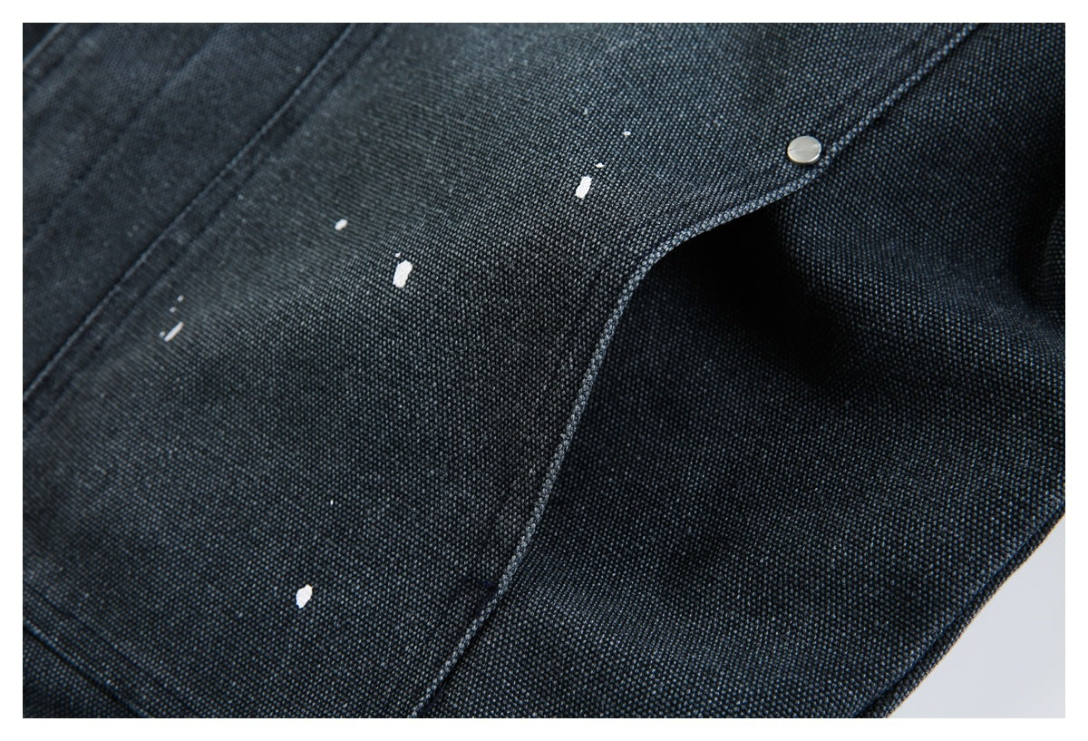 Close-up of dark indigo denim with a curved snap pocket flap and scattered white paint flecks.