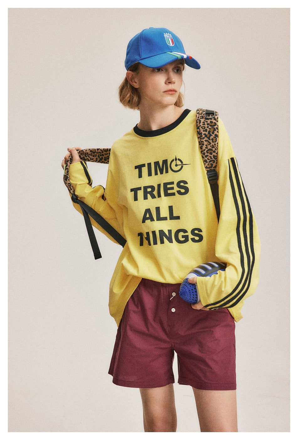 Model wearing a yellow long-sleeve shirt with black sleeve stripes and text, burgundy shorts, and a blue cap.