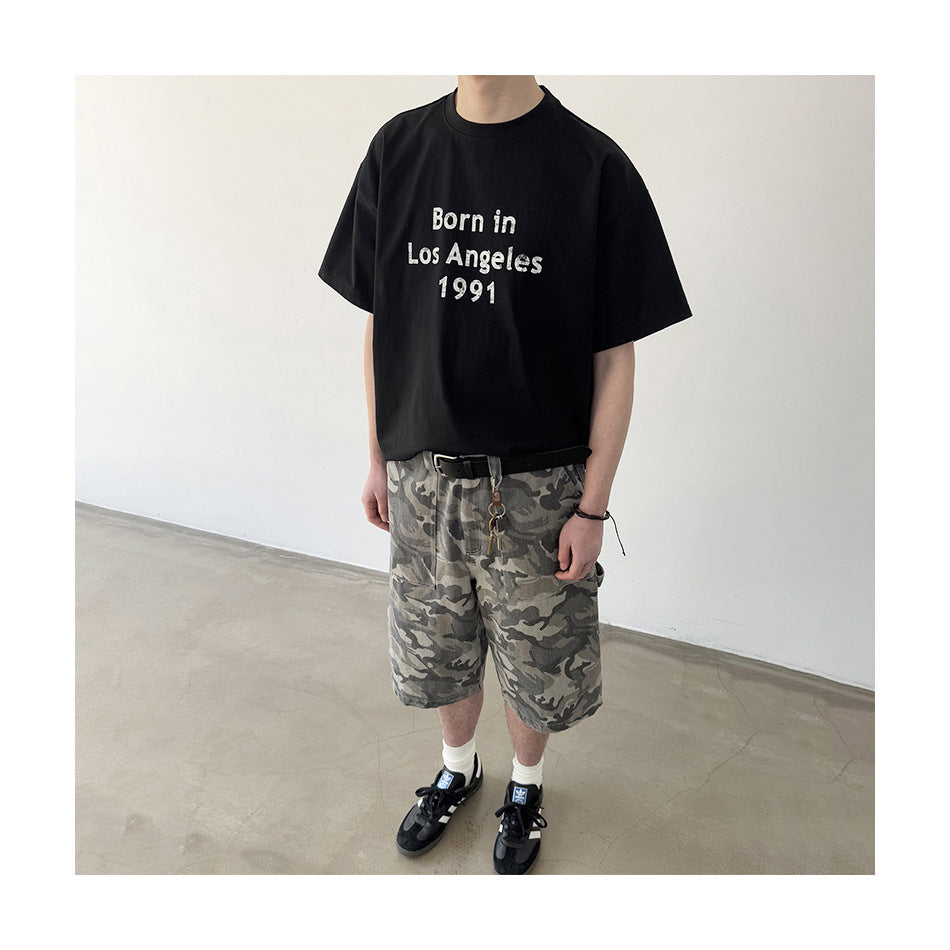 Model wearing a black “Born in Los Angeles 1991” T-shirt and gray camouflage shorts.