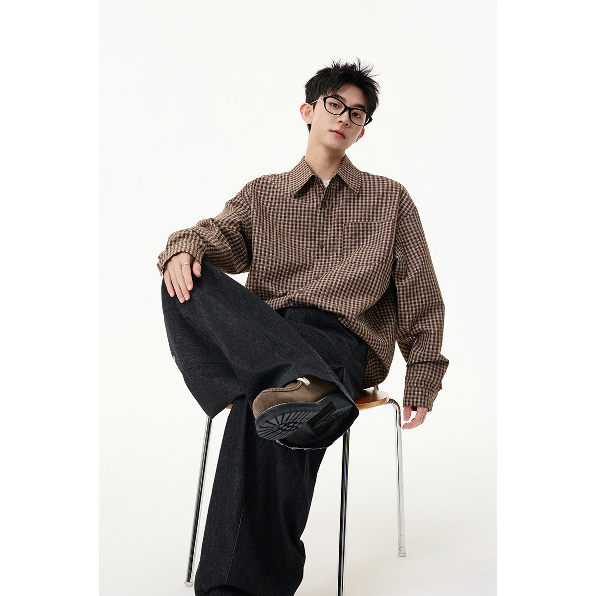 Seated model wearing a brown-and-cream checked button-up shirt with a chest pocket and dark wide-leg trousers.