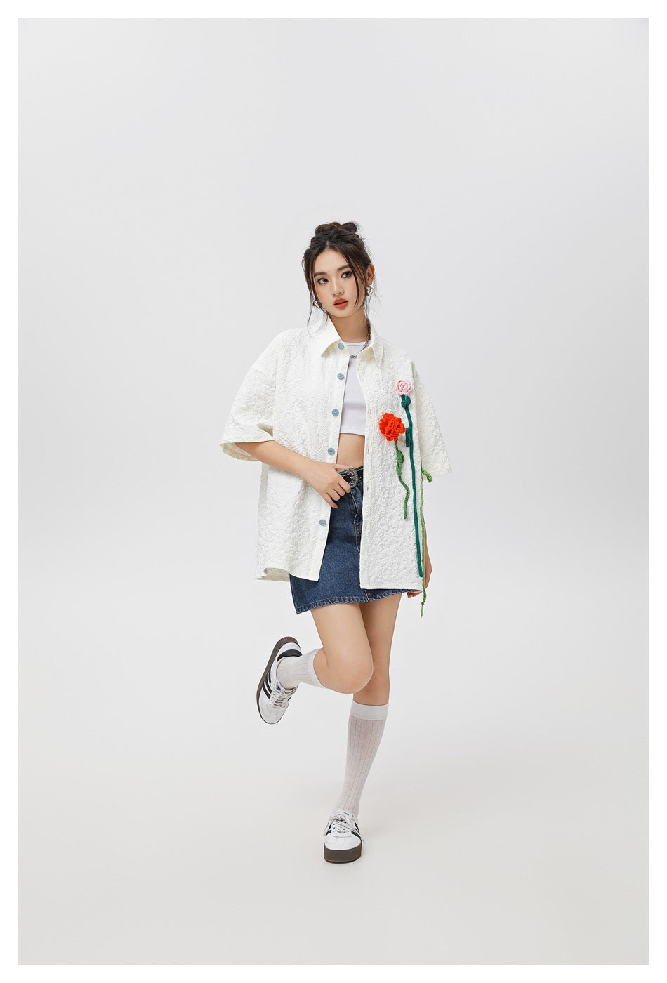 Model wearing a white textured short-sleeve button-up shirt with colorful flower appliqués and a denim skirt.
