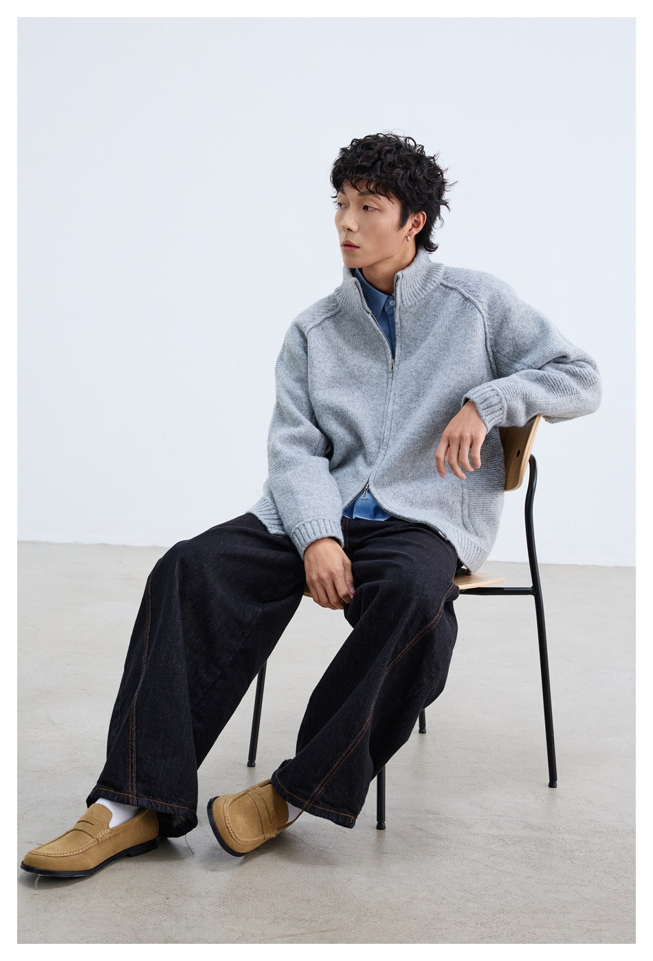 Seated model wearing a light gray zip-front knit jacket over a blue shirt with dark wide-leg jeans.