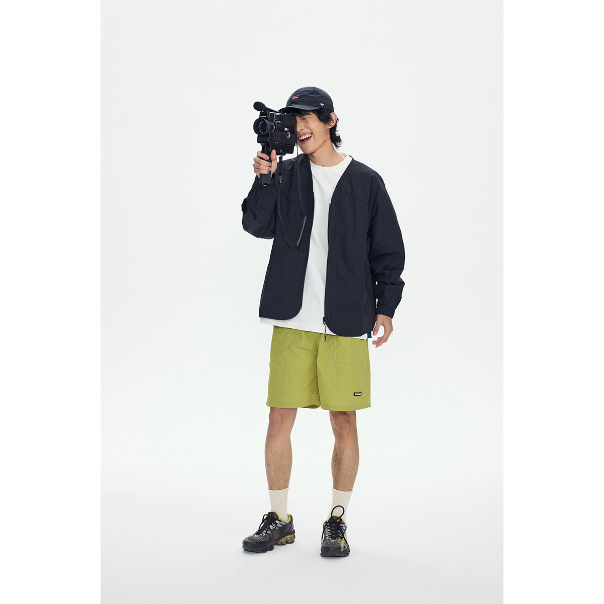 Model wearing a black open jacket, white shirt and lime green shorts while holding a video camera.