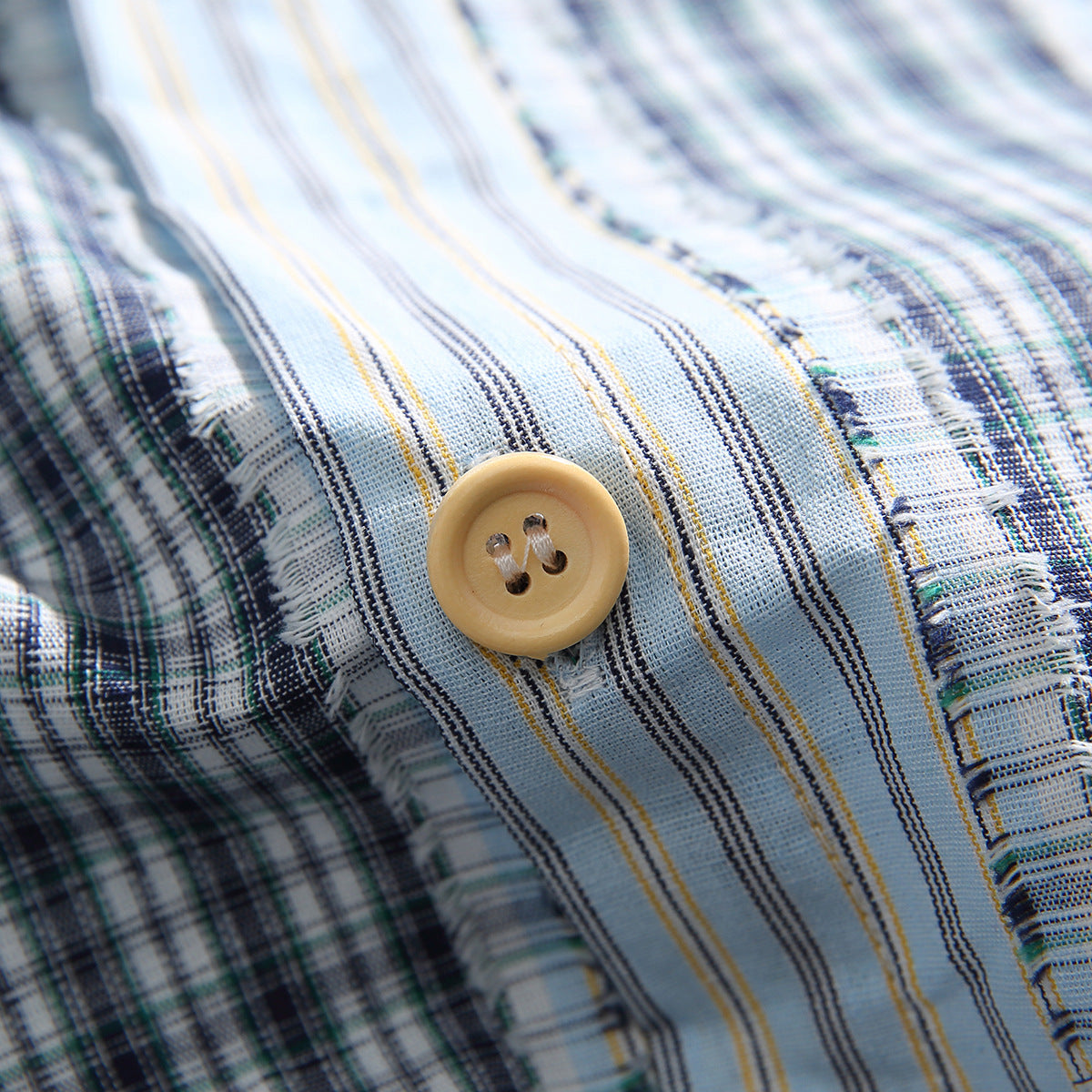 Close-up of a tan button on pale blue striped shirt fabric beside a frayed plaid panel seam.