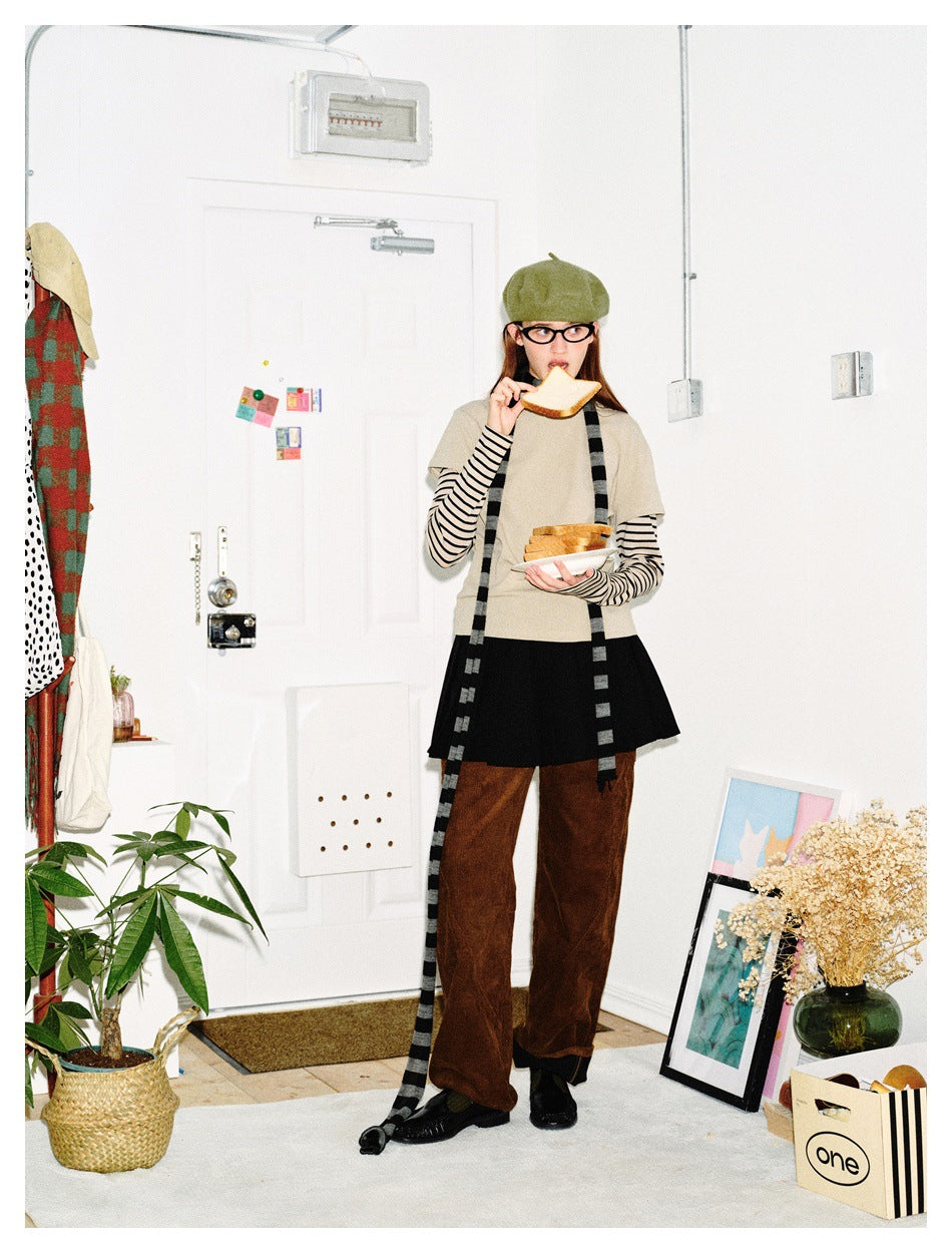 Full-length model in a beige top over striped sleeves, black skirt, brown trousers and long striped scarf, holding bread.
