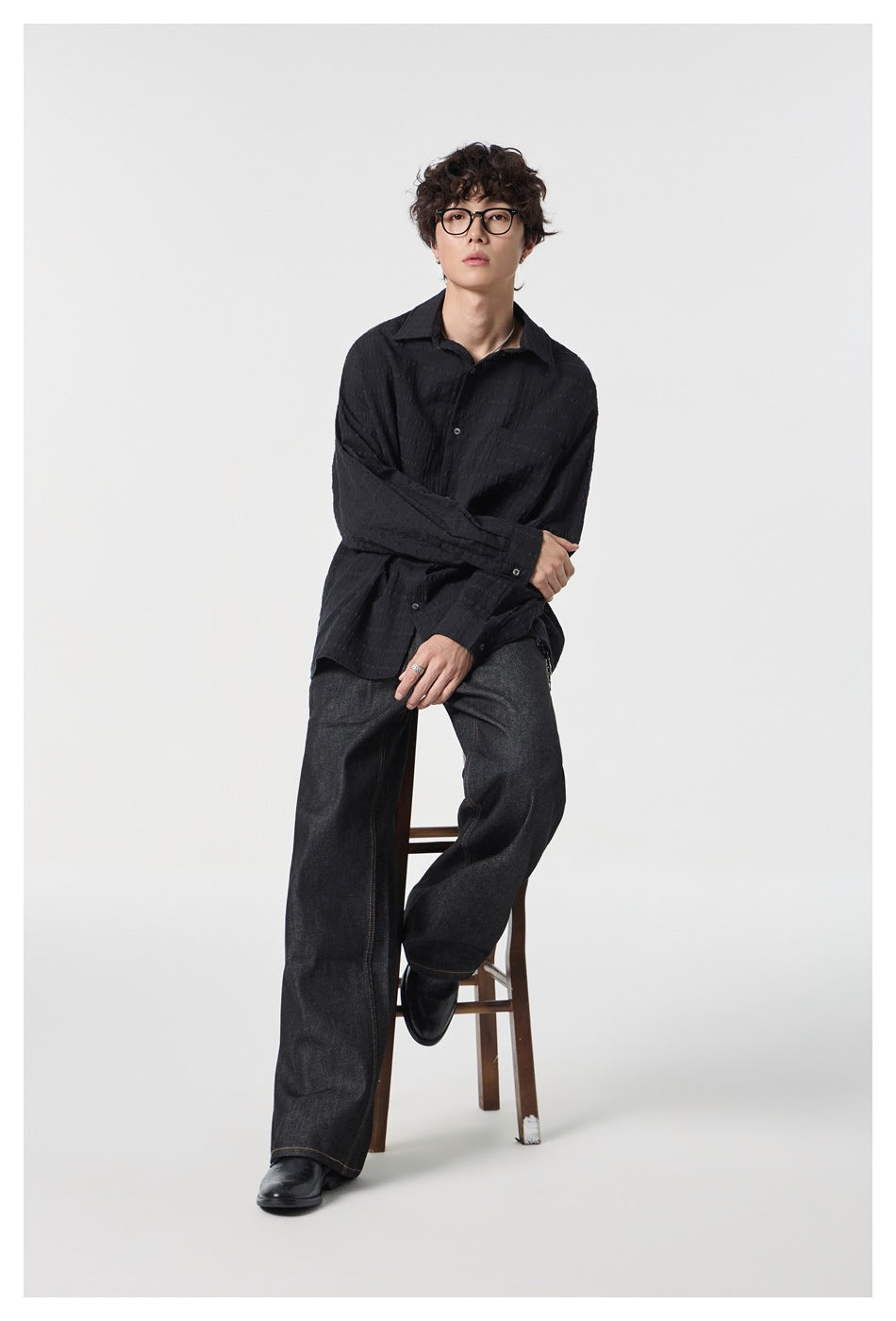 Full-length view of a model seated on a stool in a black textured shirt, dark wide-leg jeans and black leather shoes