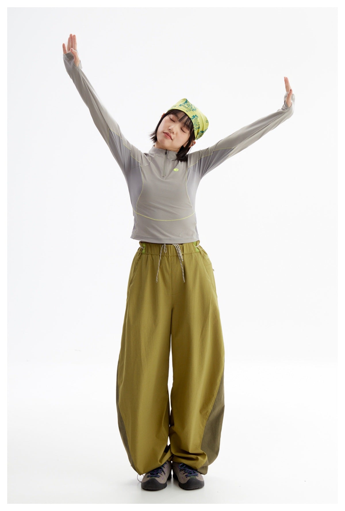 Model stretching with arms raised, wearing a gray quarter-zip top and olive wide-leg pants.