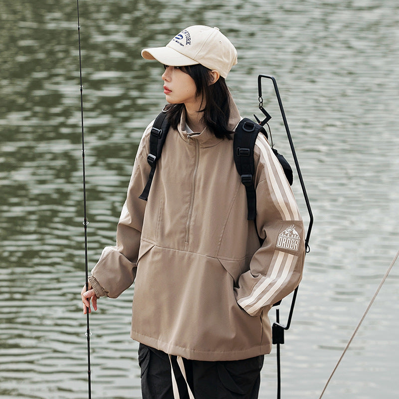 Model wearing a taupe pullover jacket with cream sleeve stripes and an embroidered sleeve graphic beside a lake.
