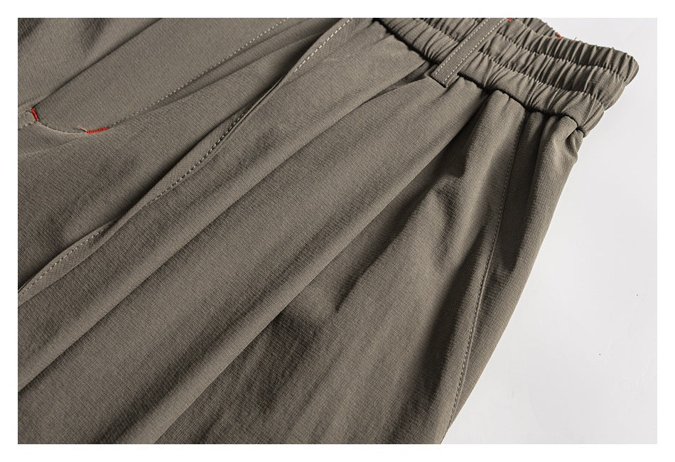 Close-up of taupe trousers showing a gathered elastic waistband, belt loop, front pocket seam, and stitching.