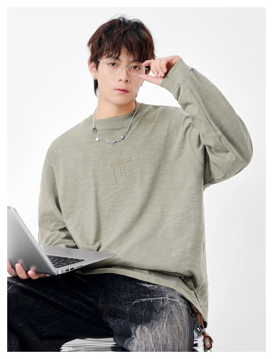 Model wearing an oversized sage-gray long-sleeve crewneck shirt with subtle chest embroidery.