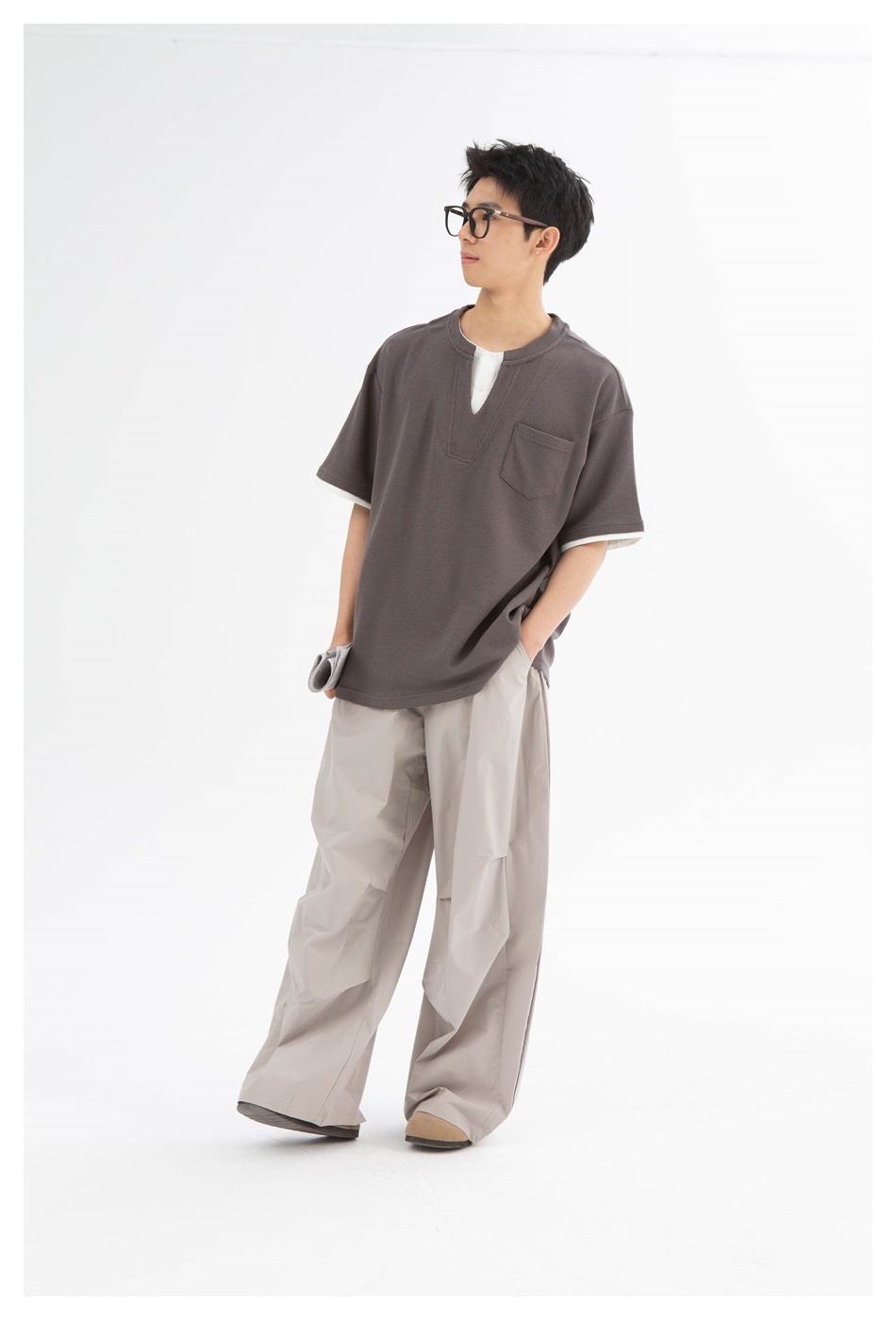 Man in a gray-brown layered short-sleeve tee with chest pocket and wide beige trousers, standing in profile