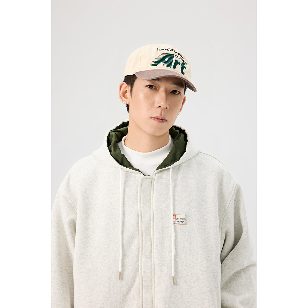 Close-up of a light gray hooded zip jacket showing dark green lining, drawstrings, and a small chest patch.
