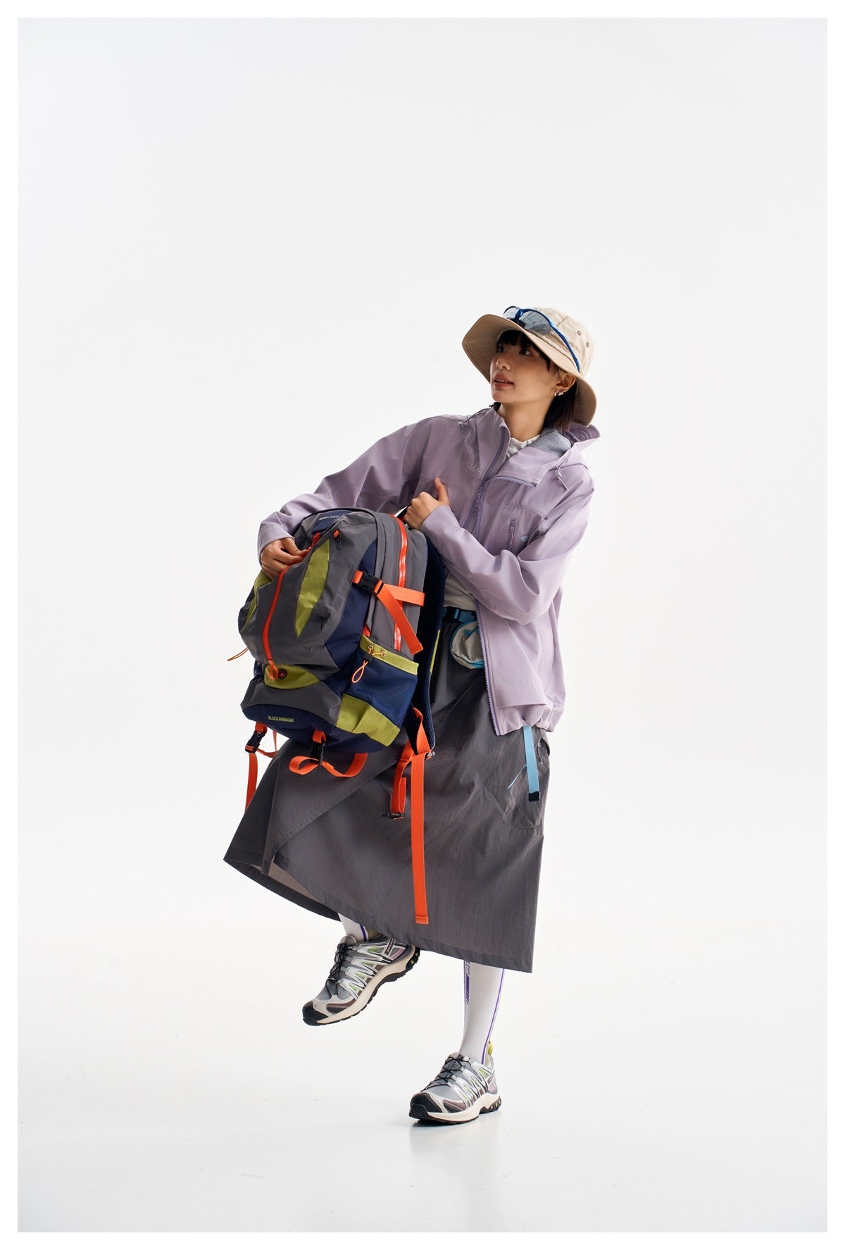 Model in a light lavender hooded jacket and dark gray skirt, holding a large multicolor hiking backpack.