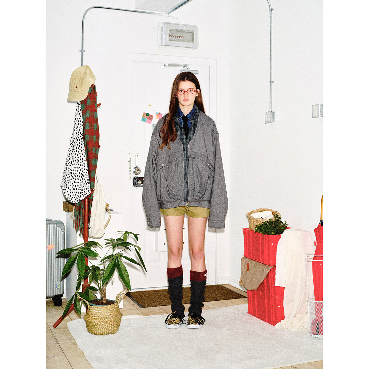 Full-length model wearing an oversized gray zip jacket over plaid layers and olive shorts, facing forward indoors.