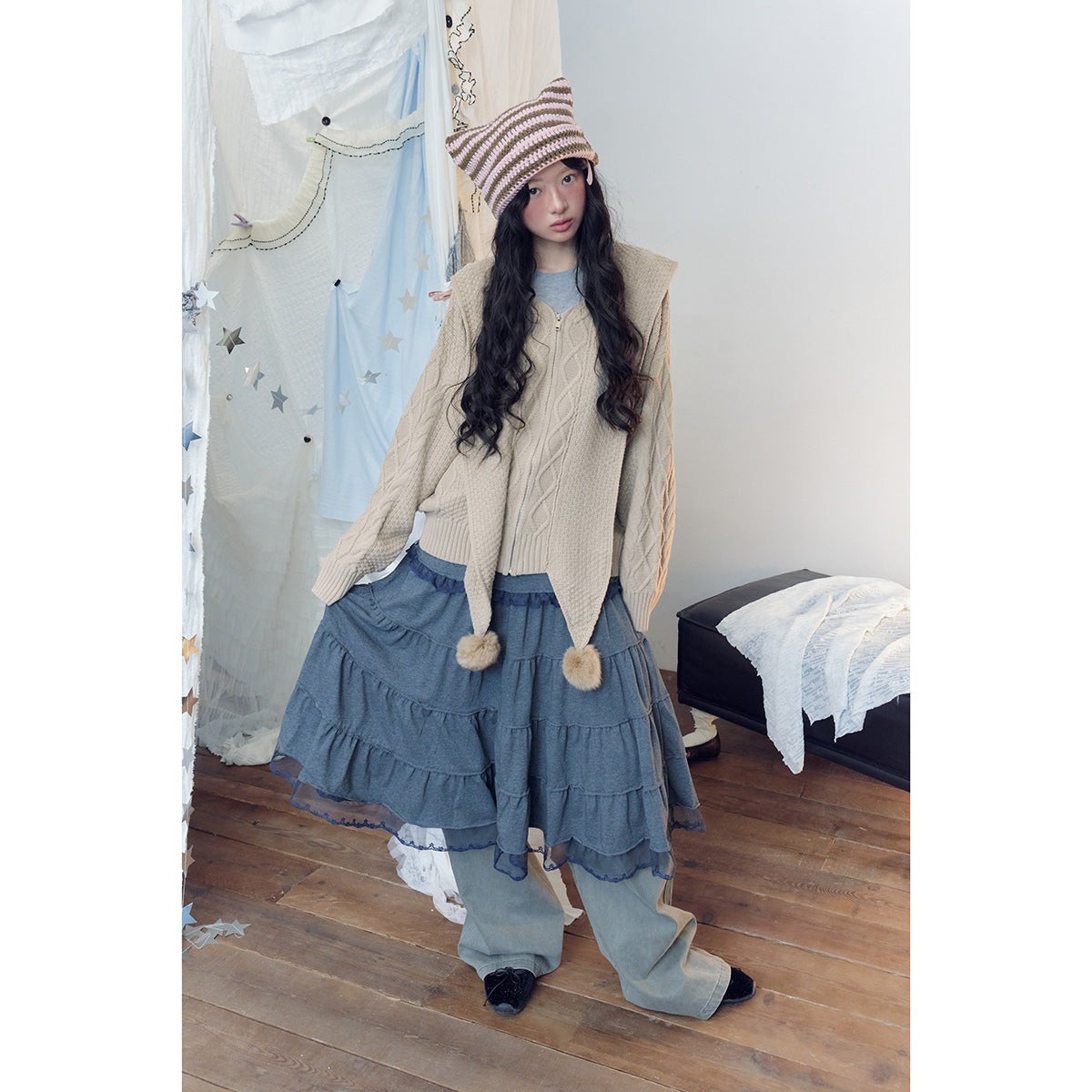 Model wears a beige cable-knit zip hoodie with pom-pom ties, a tiered blue skirt, wide-leg pants and a striped knit hat.