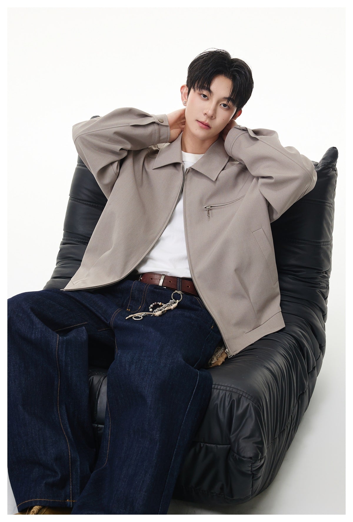 Model reclining in a black chair while wearing a light taupe zip-front jacket with a chest zipper pocket.
