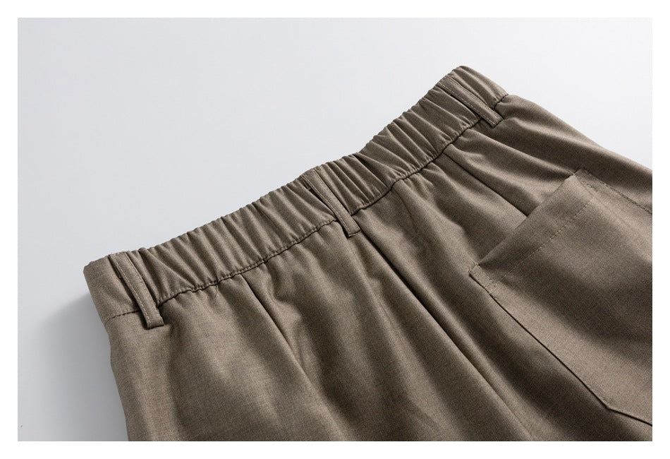 Partial front view of taupe-brown trousers with an elasticated waistband, belt loops, pleats and a pocket.