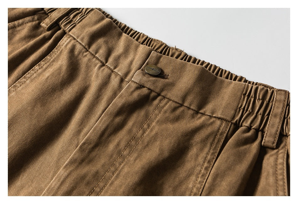 Close-up of brown trousers showing a buttoned waistband, elasticated sides, belt loops and front seams.