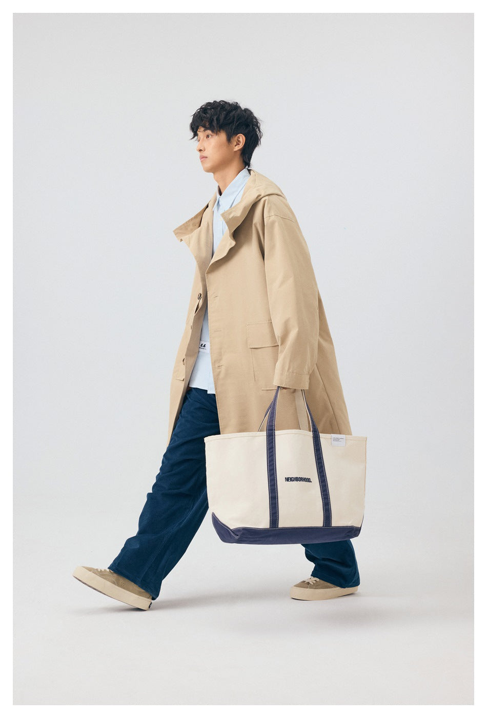 Model wearing a long beige hooded coat over a blue shirt and navy trousers, carrying a cream-and-navy tote.