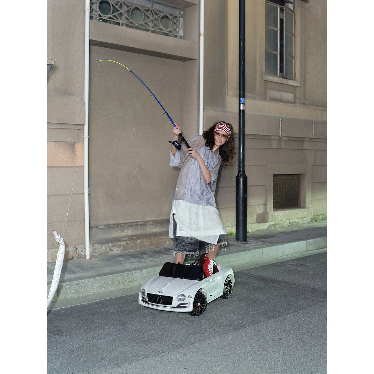 Model wearing a pale gray layered outfit while holding a fishing rod on a city street.