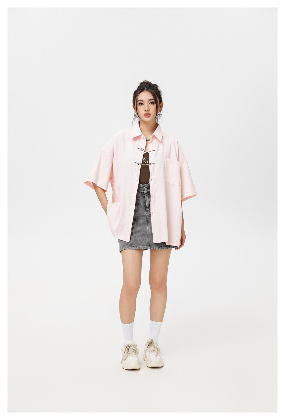 Model wearing a pale pink oversized short-sleeve shirt with a chest pocket and decorative clasps, layered over a top and gray