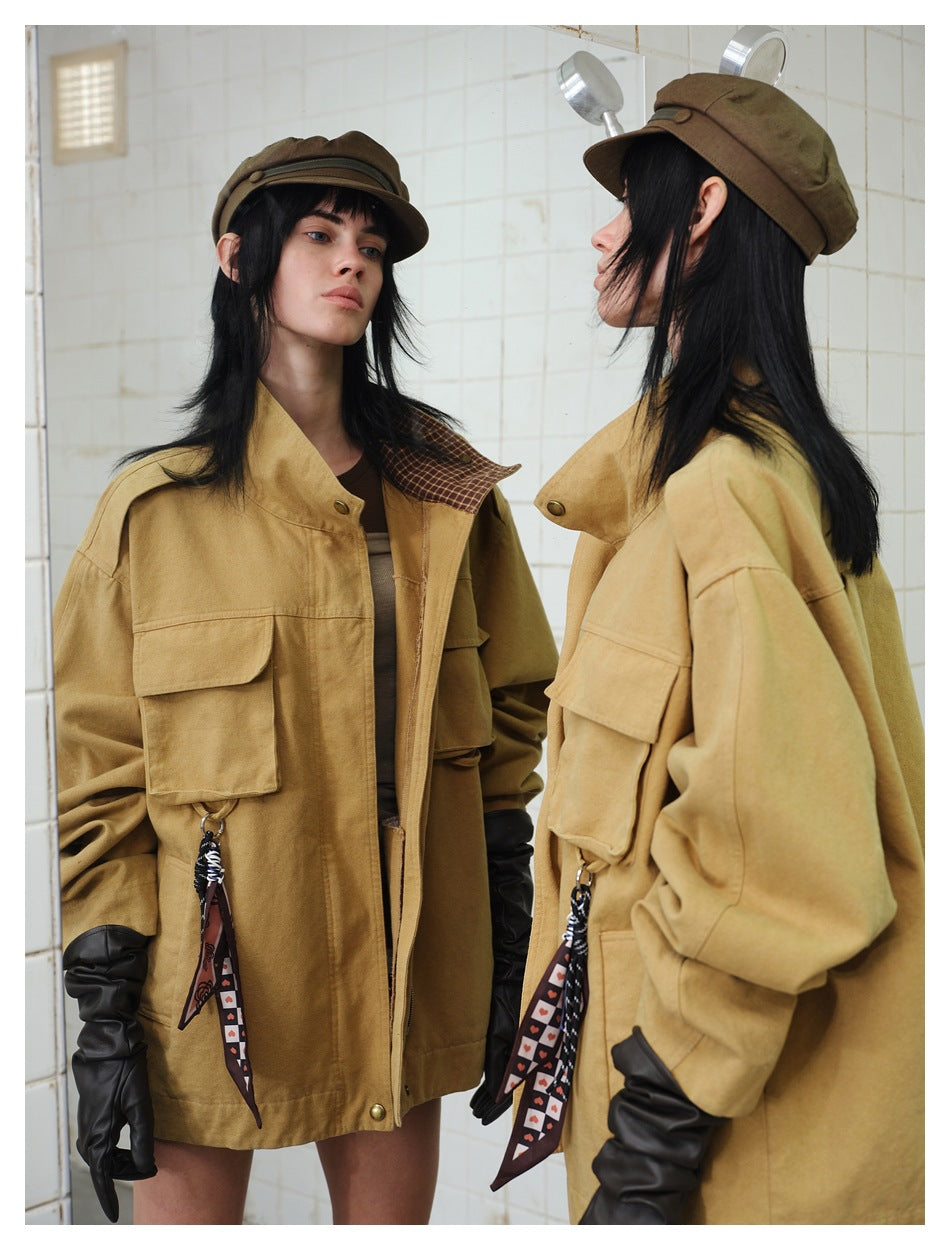 Model in brown jacket with scarf charm, shown facing a mirror in a tiled room with a cap and black gloves