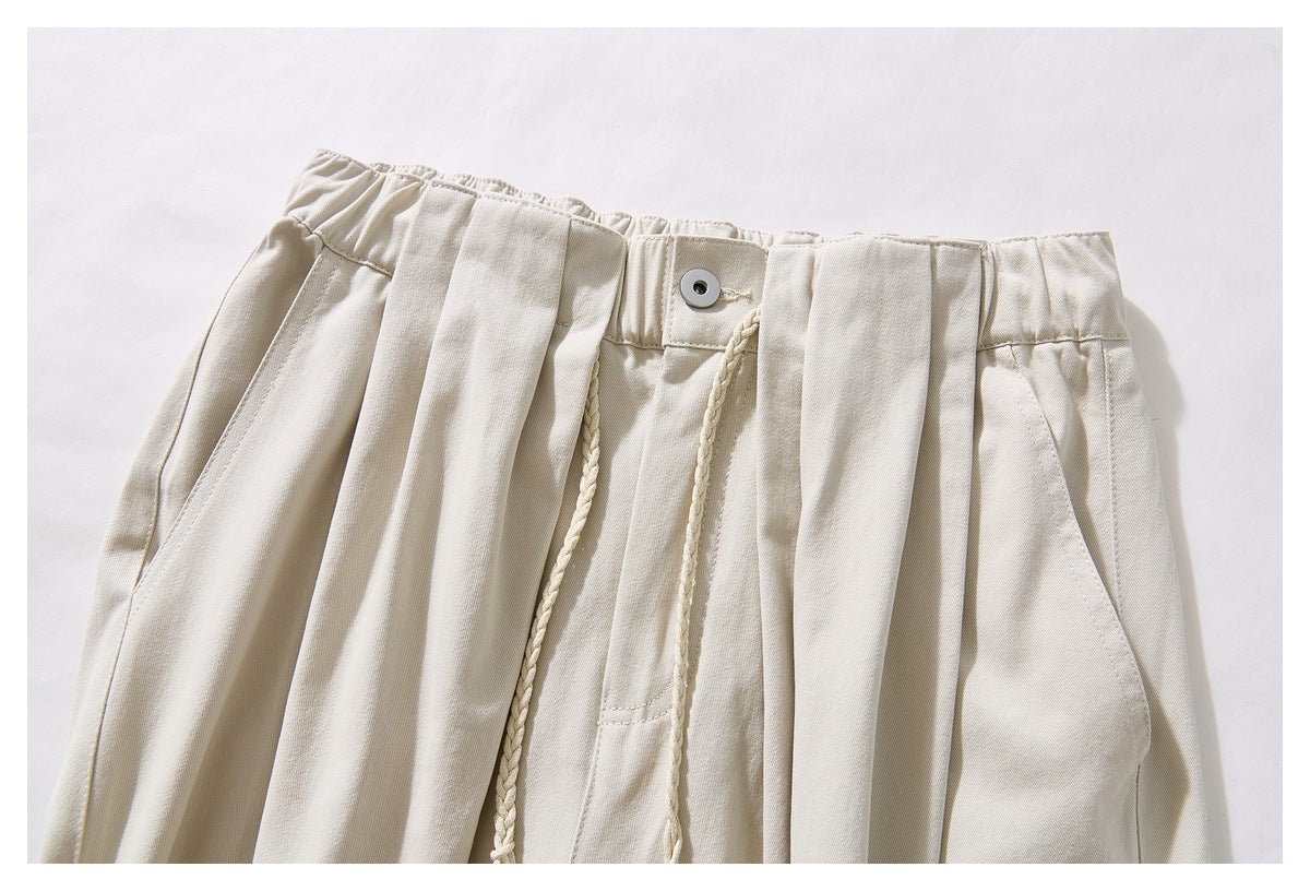 Close-up of cream trousers showing a gathered waistband, front button, pleats, and braided drawstrings.