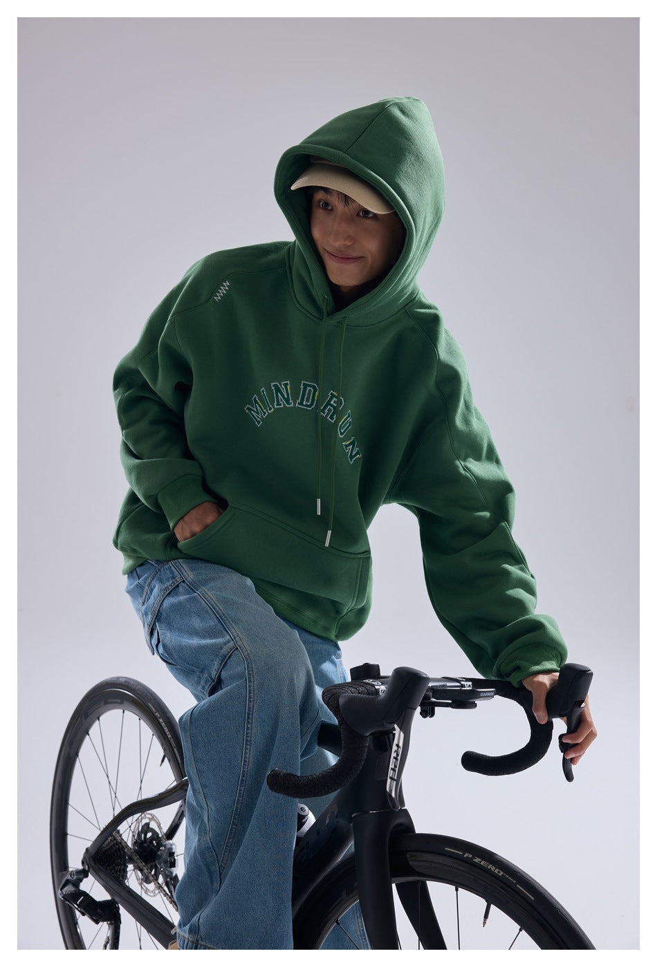 Model wearing a dark green hoodie with the hood up, arched chest lettering and a kangaroo pocket while leaning over a