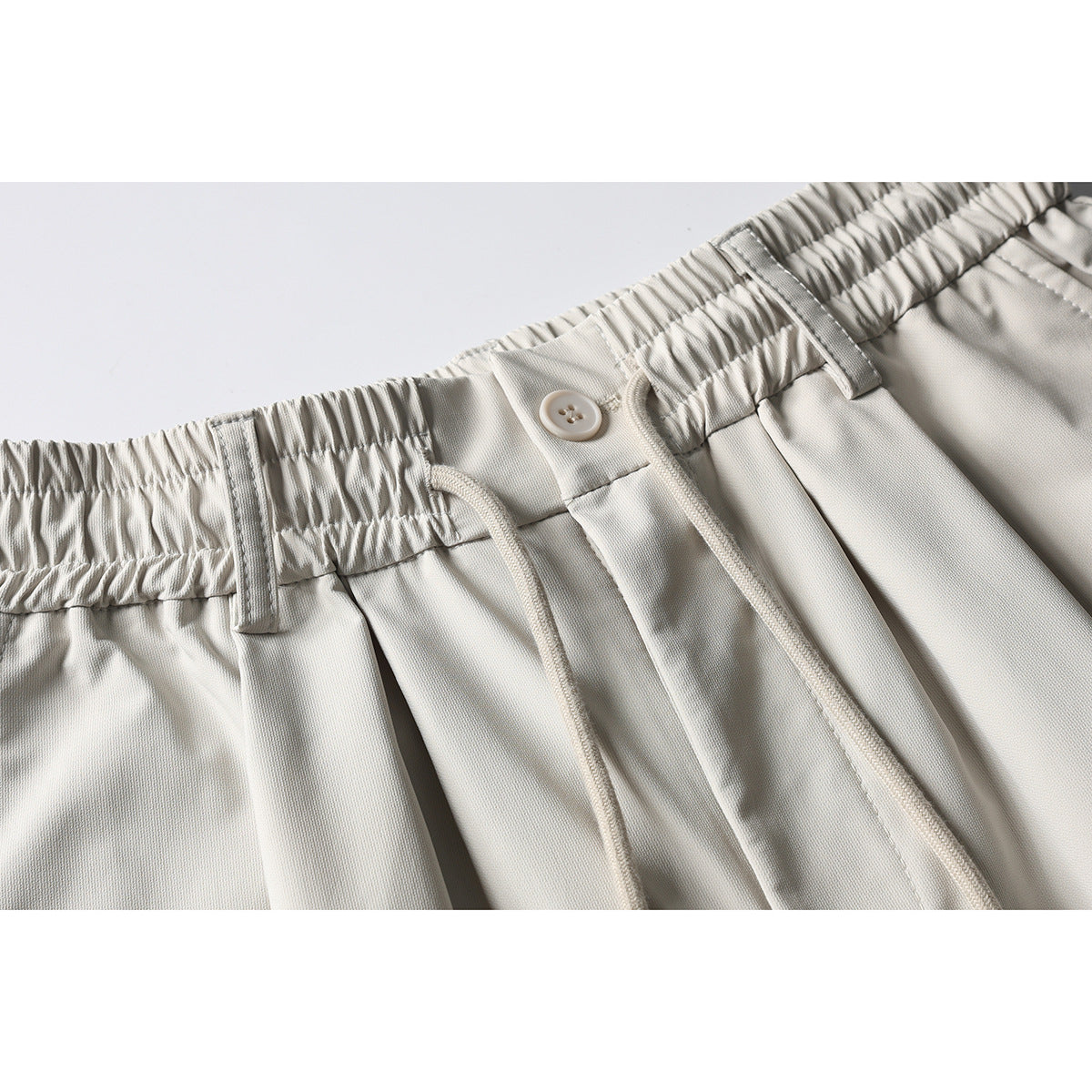 Close-up of light beige shorts showing the gathered elastic waistband, button, belt loops, pleats and drawstrings.