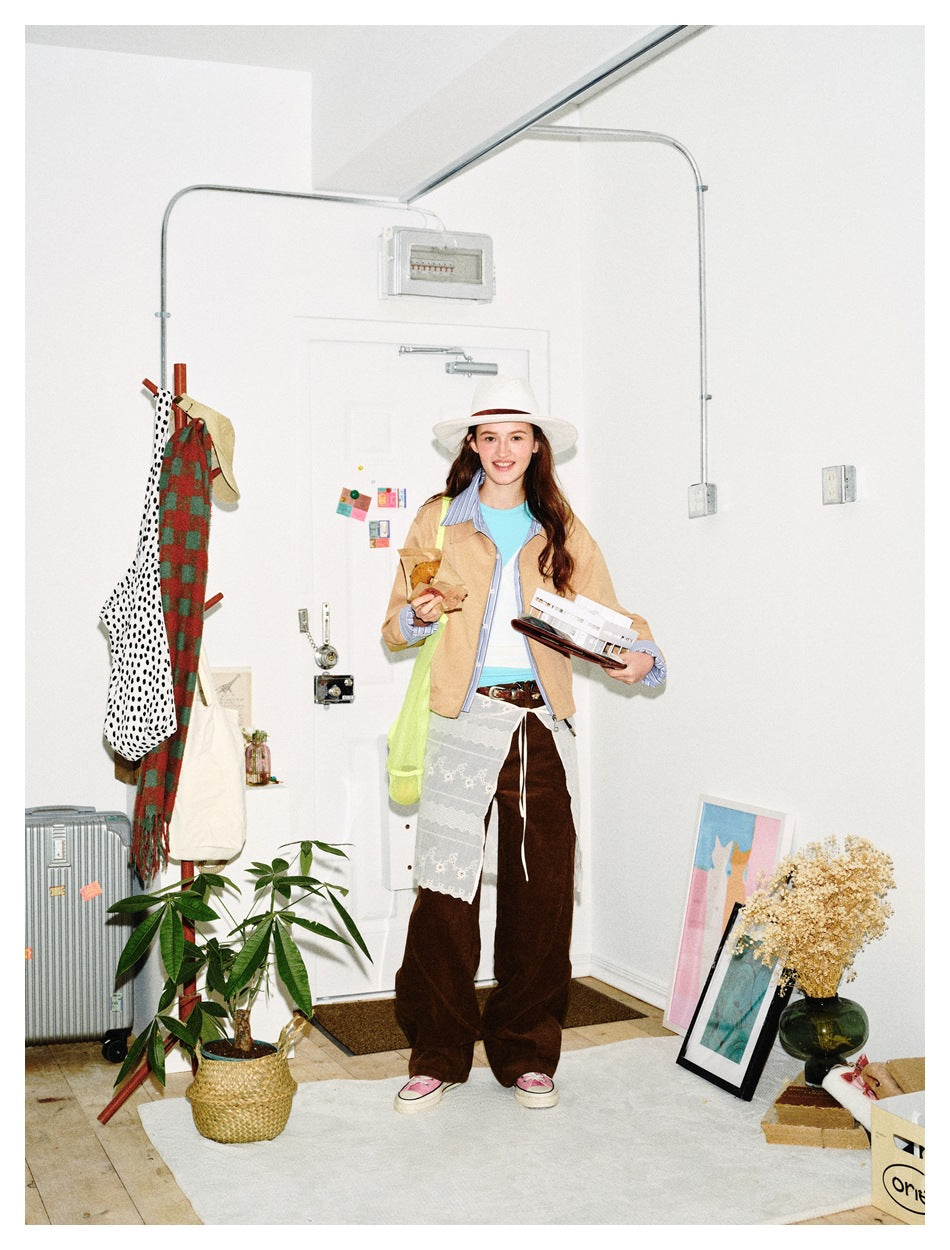 Woman in a khaki zip jacket with striped shirt layer, hat, brown trousers and lace skirt layer in a styled room