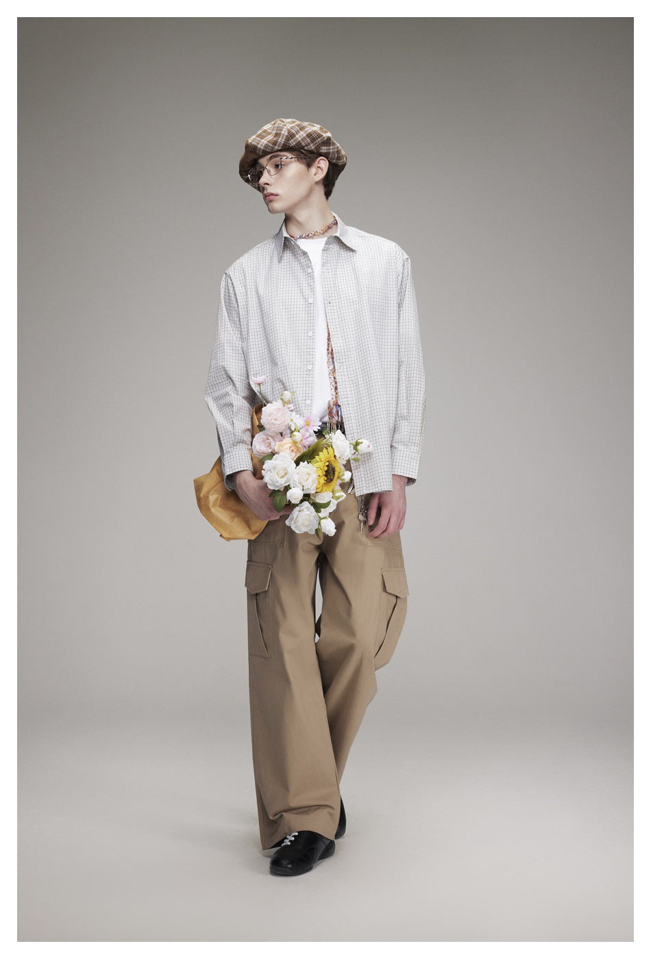 Model wearing an oversized light gray checked shirt and tan cargo trousers, holding a bouquet of flowers.