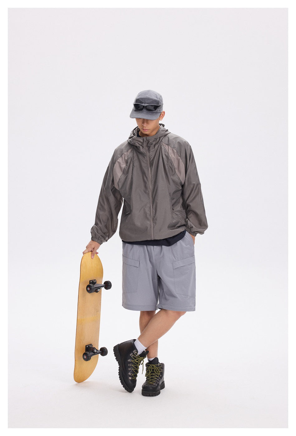 Person wearing a dark gray hooded zip jacket with gray cargo shorts, cap and boots, holding a skateboard