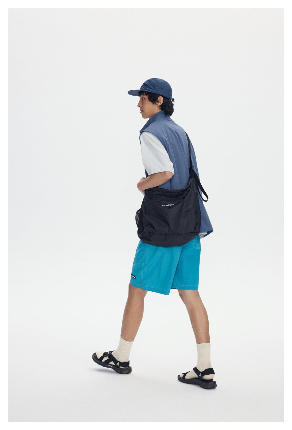 Side-rear view of a model walking in a slate-blue vest and turquoise shorts with a black shoulder bag.