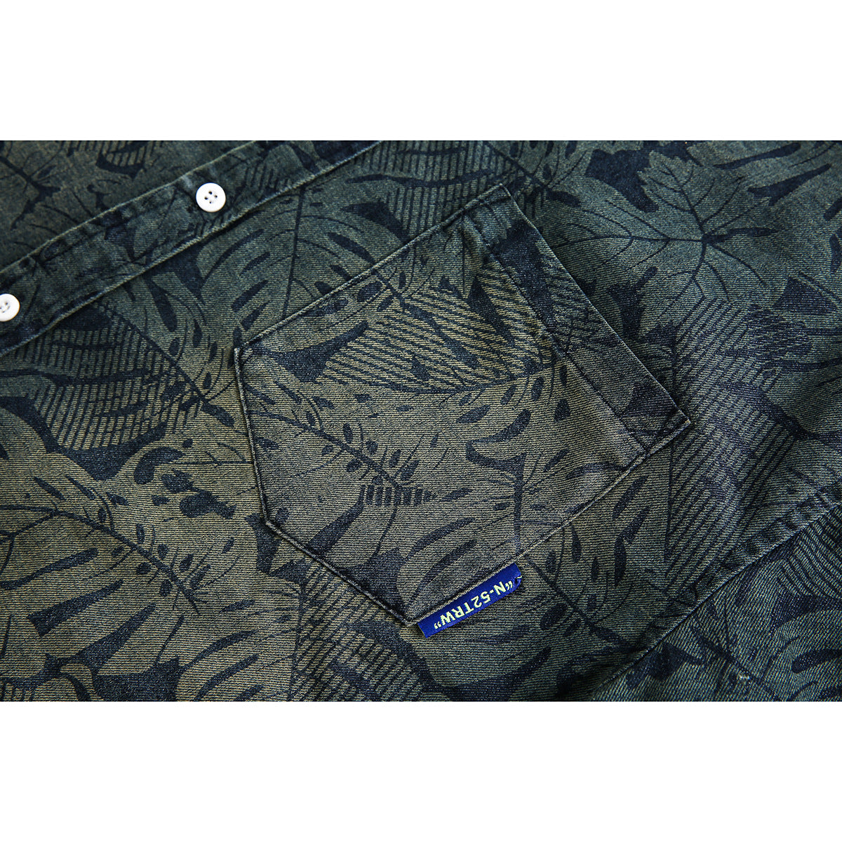 Close-up of a buttoned chest pocket on a dark blue-gray shirt with an abstract botanical print and small blue tag.