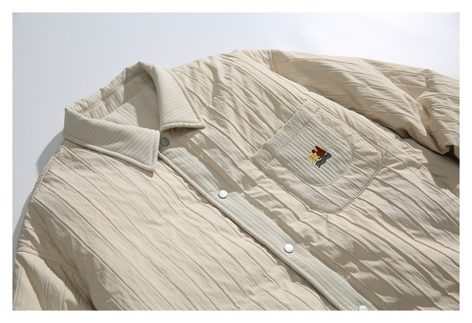 Angled close view of an ivory quilted jacket front, showing its snap buttons, pointed collar, and embroidered chest pocket.