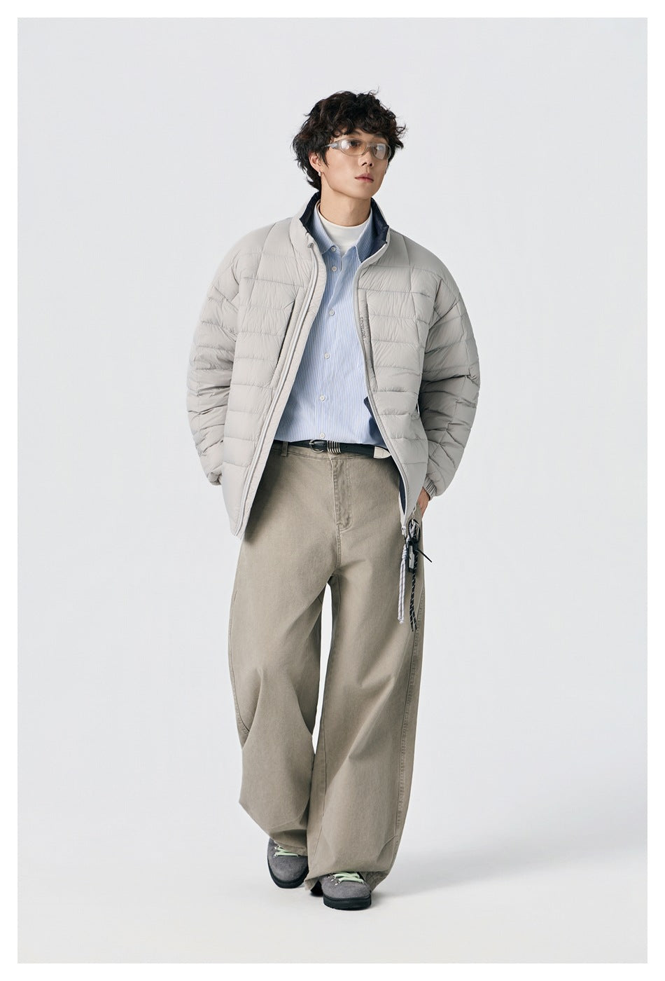 Model walking in an open light gray quilted jacket over a blue striped shirt and loose beige trousers.