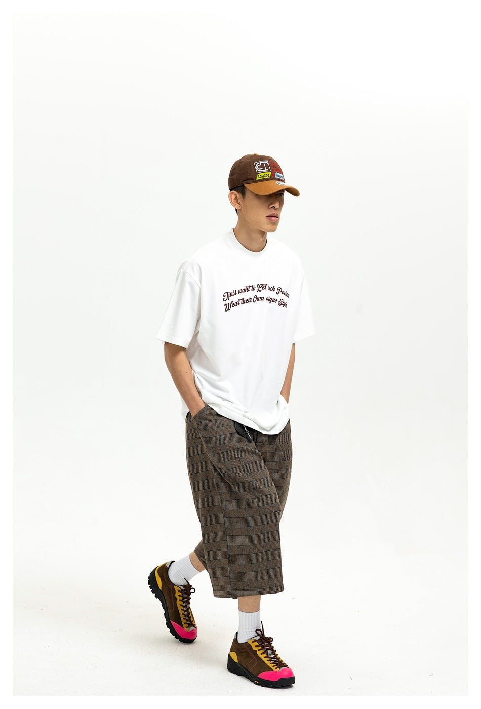 Man walking in a white slogan T-shirt with brown plaid shorts, a cap and colorful sneakers, three-quarter view