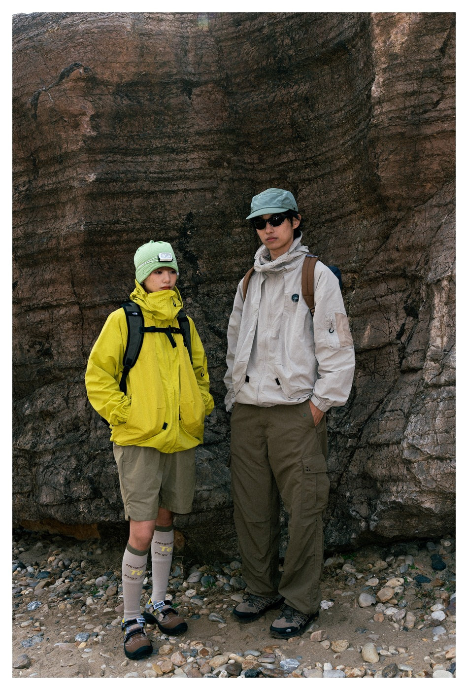 Two models outdoors wearing bright yellow and light beige hooded jackets with casual cargo bottoms.