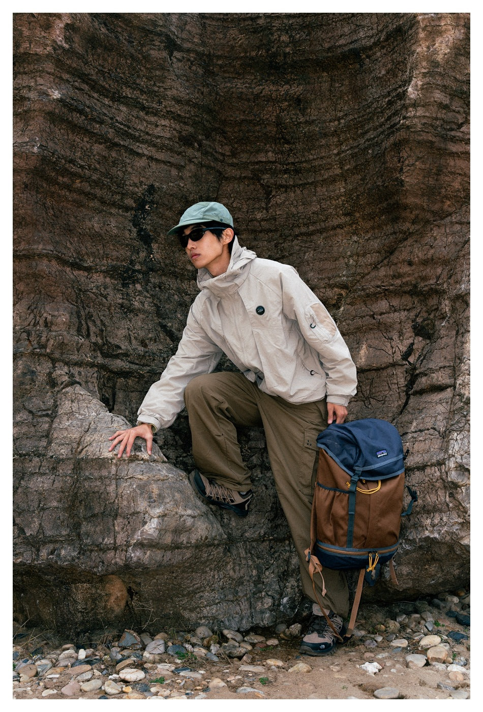A model wears a light beige hooded jacket and olive trousers while climbing beside a rock wall.