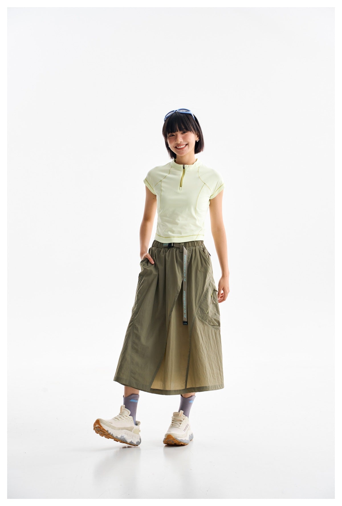 Model wearing a pale yellow zip-neck athletic top and long olive wrap skirt in a white studio.