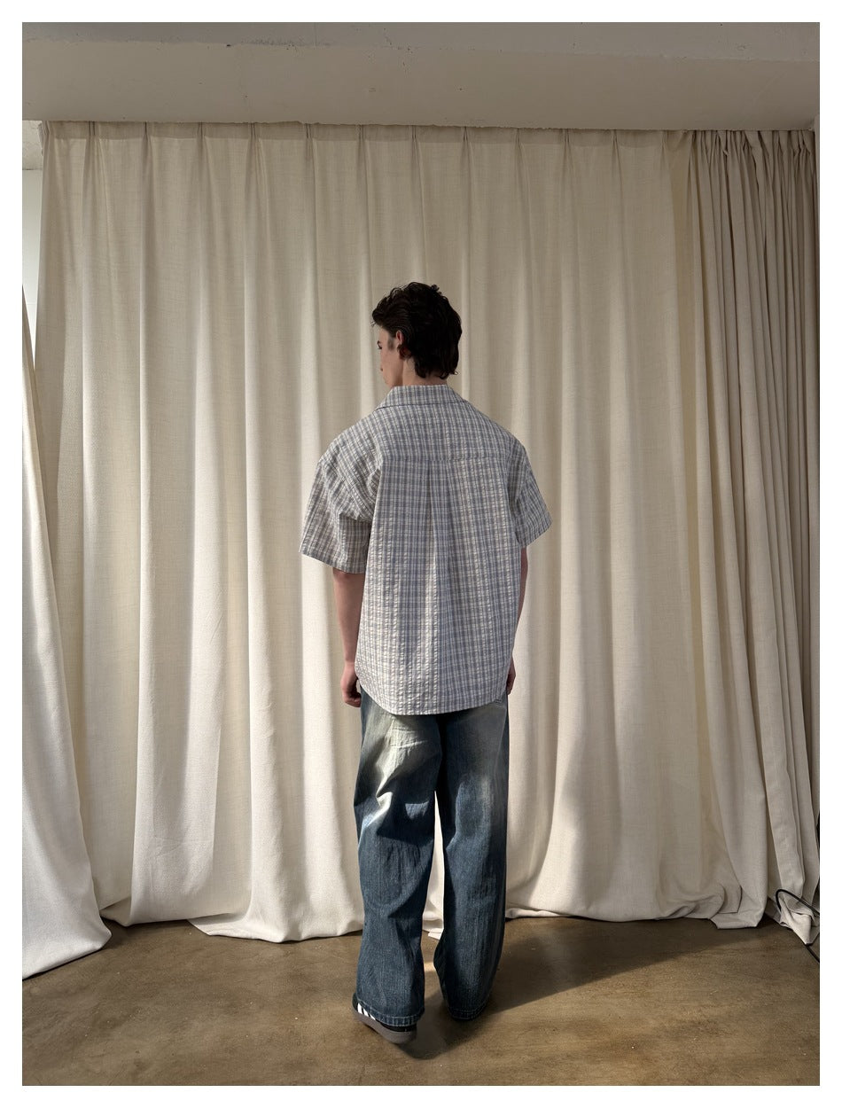 Back view of a man in a blue-gray plaid short-sleeve shirt and loose dark jeans, framed by a white border.