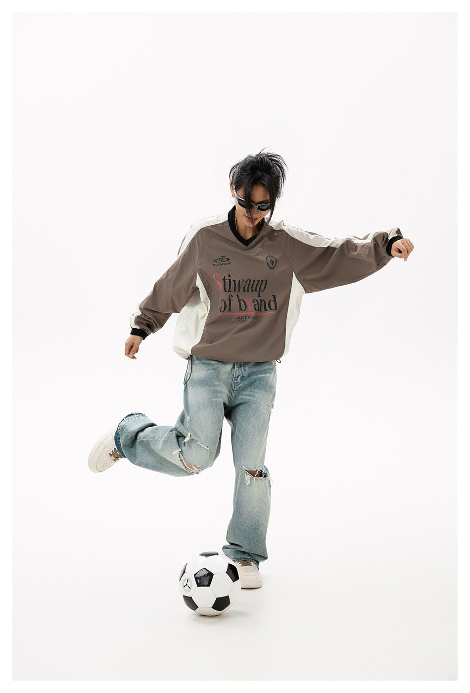 Model wearing a taupe and cream V-neck pullover with black trim, posed with a soccer ball.