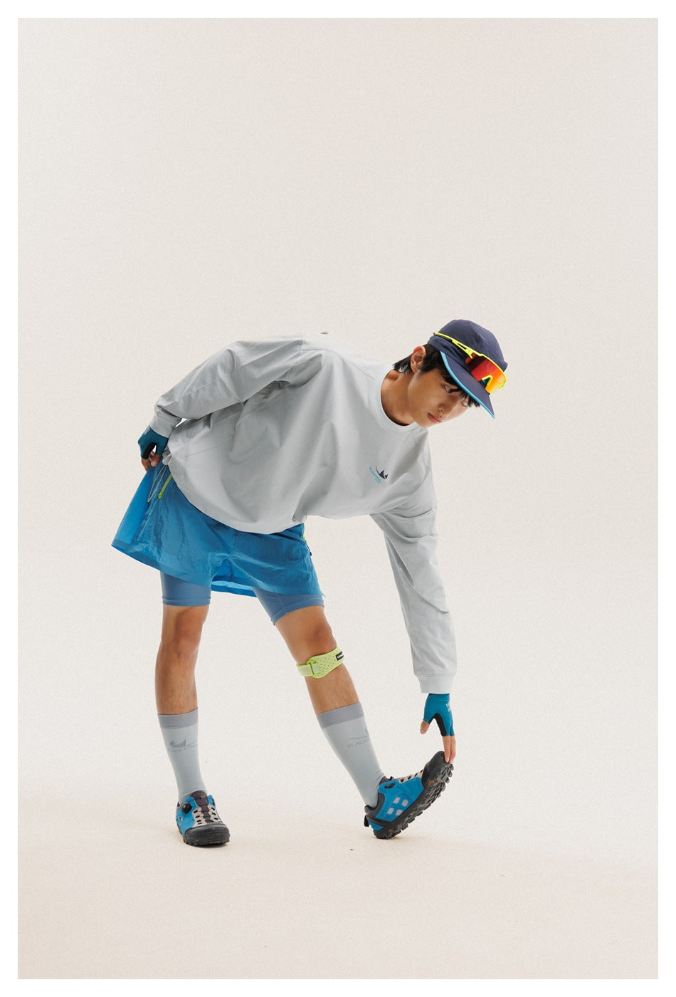 Model bending forward in a light gray long-sleeve shirt and bright blue shorts.