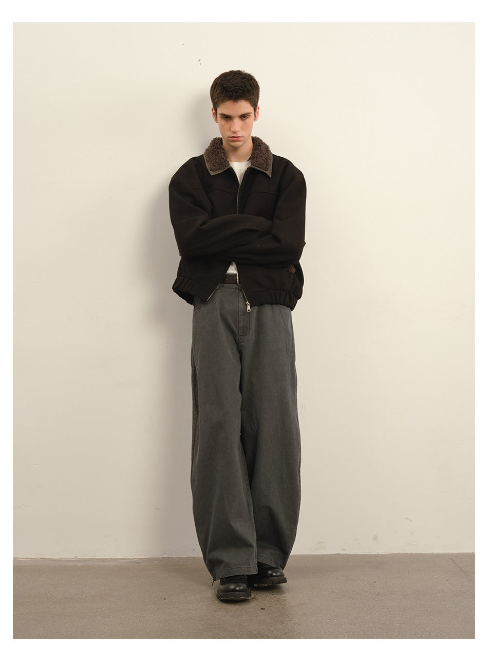 Model leaning on a wall with arms crossed, wearing a dark cropped jacket with fleece-like collar and wide gray pants