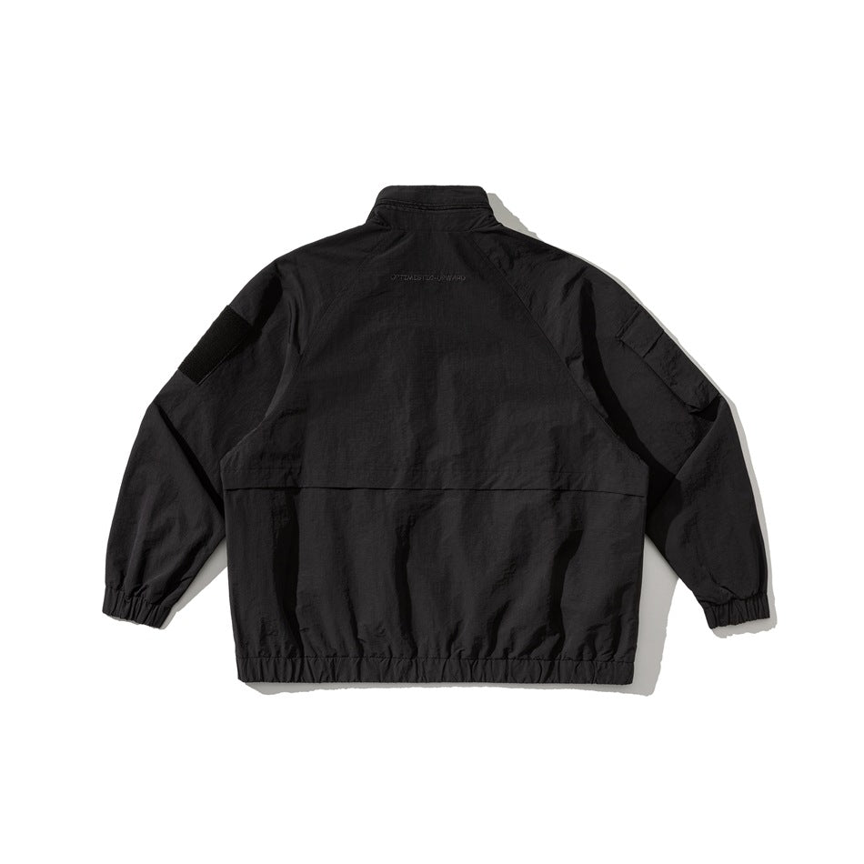 Back view of a black utility jacket with a high collar, raglan sleeves, black upper-sleeve patches, and an elastic hem.
