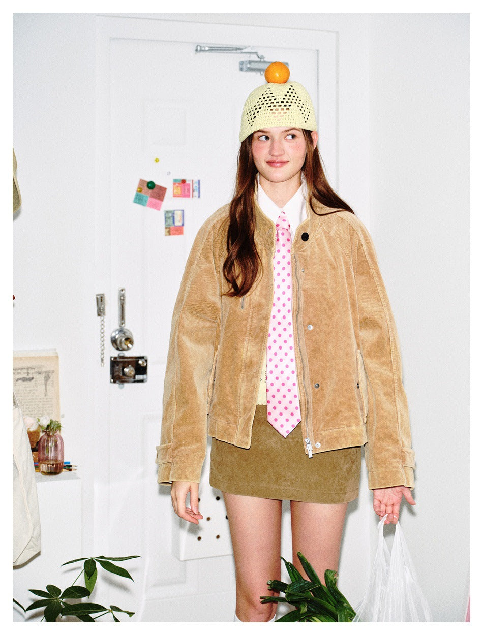 Model wearing a tan textured jacket over a white shirt and pink polka-dot tie, with an olive mini skirt and cream knit cap.