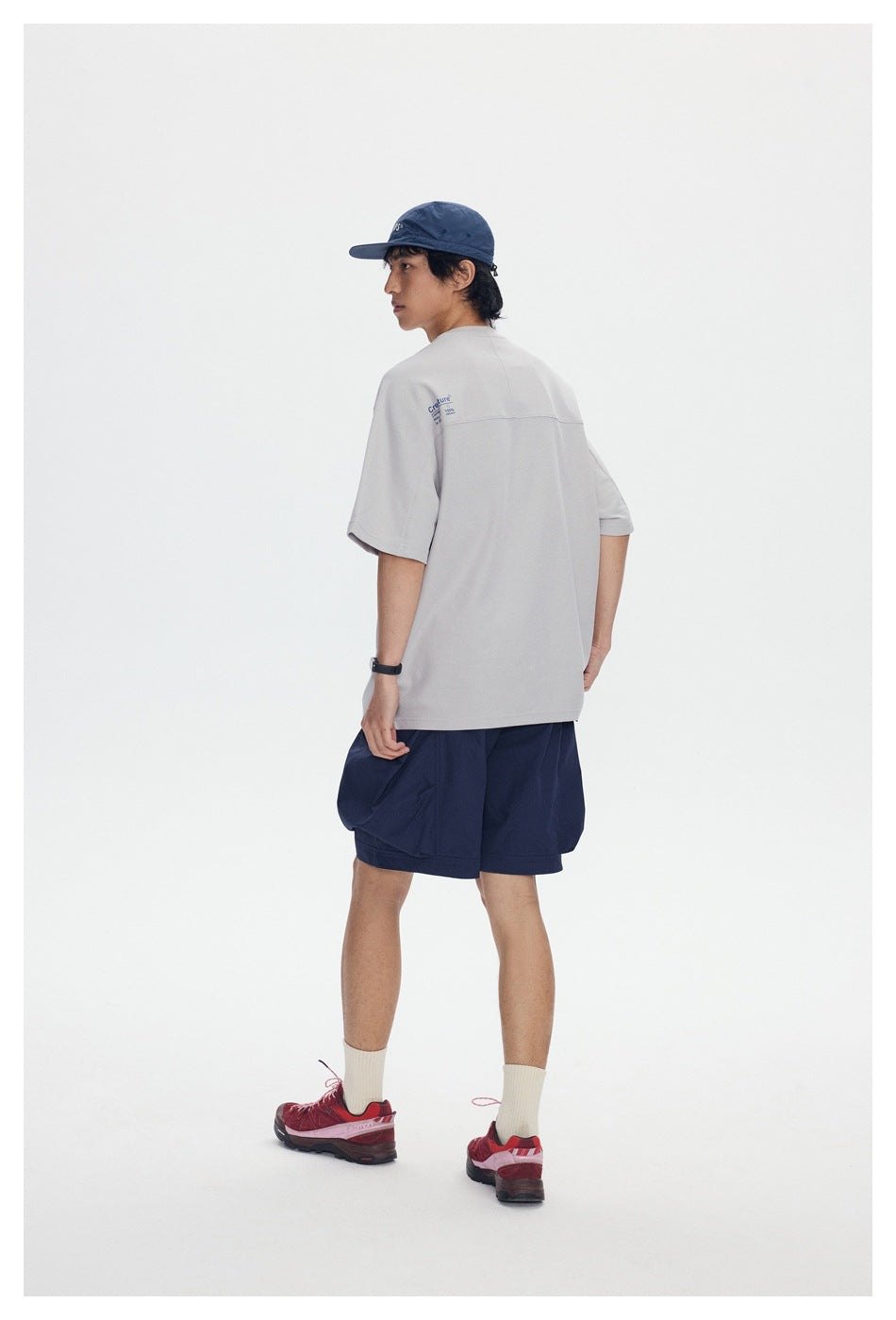 Model seen from the back in light gray T-shirt with upper-back seam and small blue print, navy shorts and red sneakers